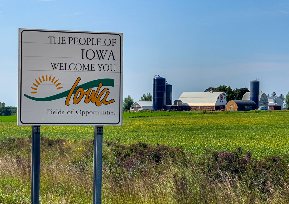 Willkommensschild des US-Bundesstaates Iowa mit der Aufschrift „The People of Iowa Welcome You – Fields of Opportunities“. Im Hintergrund liegen grüne Felder und ein landwirtschaftlicher Betrieb mit Scheunen und Silos unter blauem Himmel.