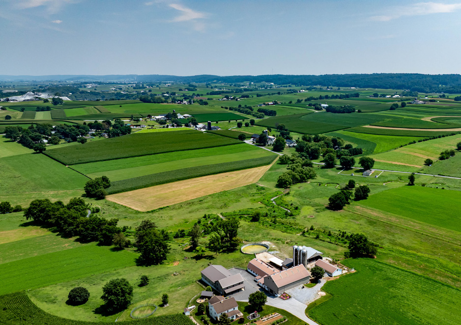 Luftaufnahme einer weitläufigen ländlichen Landschaft mit unterschiedlich bewirtschafteten grünen Feldern, Baumgruppen und verstreuten Bauernhöfen. Im Vordergrund liegt ein größerer landwirtschaftlicher Betrieb mit mehreren Gebäuden und Silos.