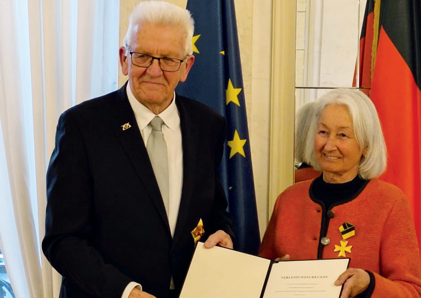 Ministerpräsident Winfried Kretschmann und Verena Schiltenwolf halten bei der Verleihung des Verdienstordens des Landes Baden-Württemberg am 27. März 2026 gemeinsam die Urkunde. Im Hintergrund sind die Flaggen der Europäischen Union und Deutschlands zu sehen.